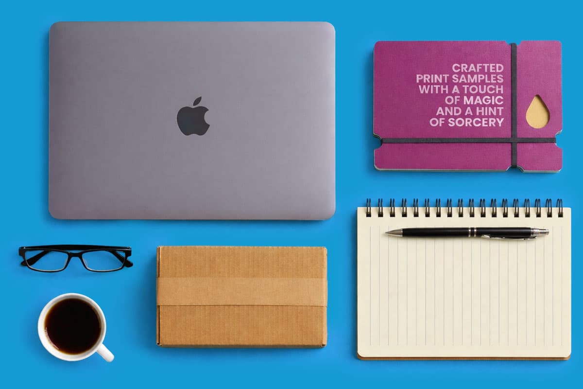 Laptop, notebook with pen, glasses, coffee, box, and purple print samples on blue background.