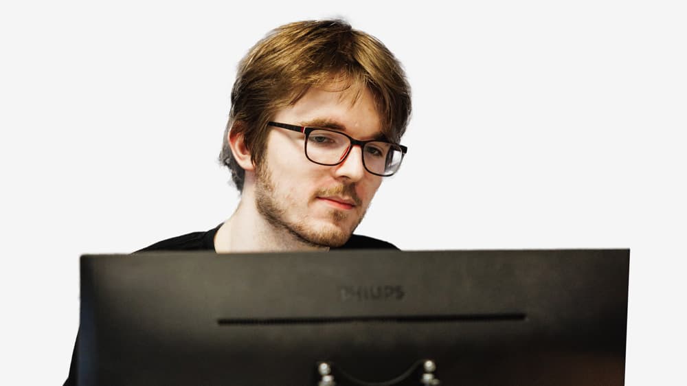 A man with glasses looks at a computer monitor against a plain white background.