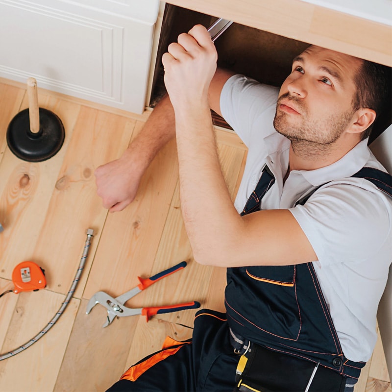 Plumber repairing pipes under a sink with tools and a plunger on the wooden floor nearby.